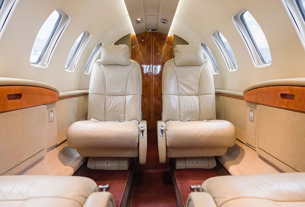 The aircraft cabin shows two beige leather seats facing forward, with windows on either side and a wooden bulkhead in the background.