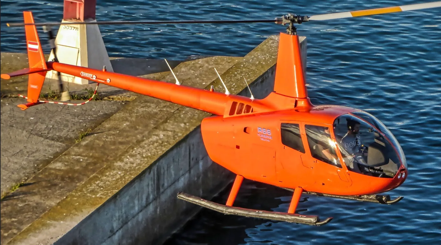 An orange Robinson R66 helicopter is shown in flight near the water.