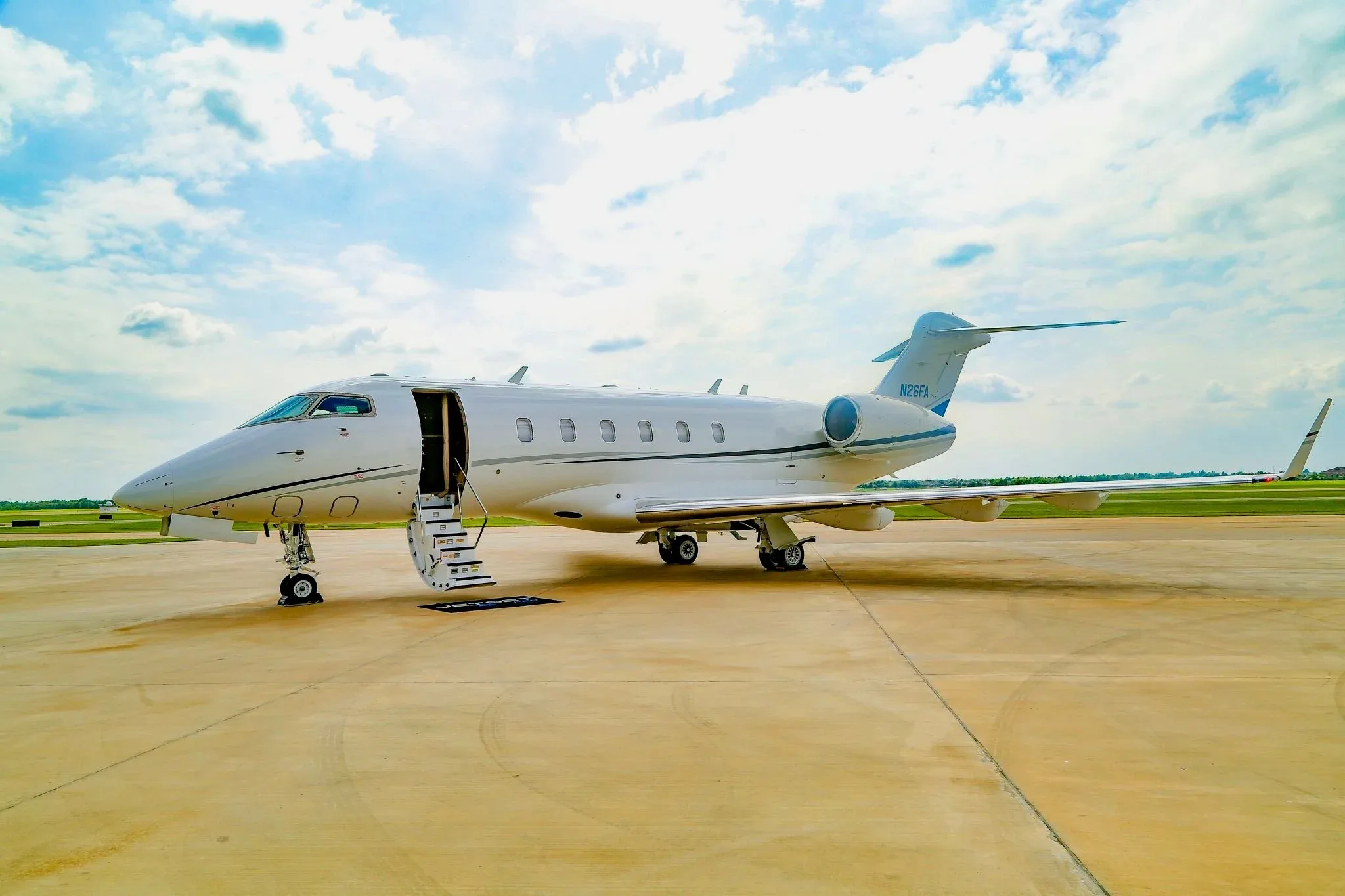 A white Bombardier Challenger 350 business jet, registration N26FA, is parked on a tarmac with its main door open and stairs extended.