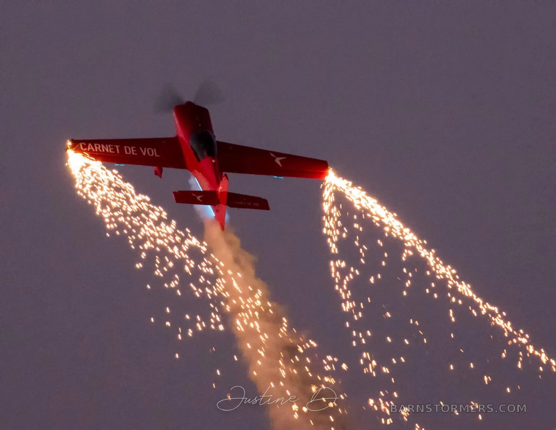 A red aerobatic aircraft with 'CARNET DE VOL' on its wing is flying upside down, trailing sparks from pyrotechnics against a dark sky.
