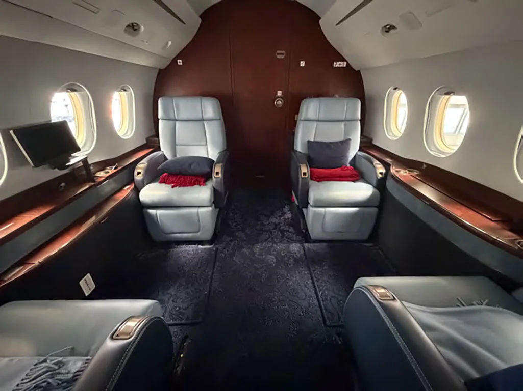 A view inside the business jet cabin showing two pairs of light blue leather seats with red and dark blue throws, and dark wood paneling at the rear.