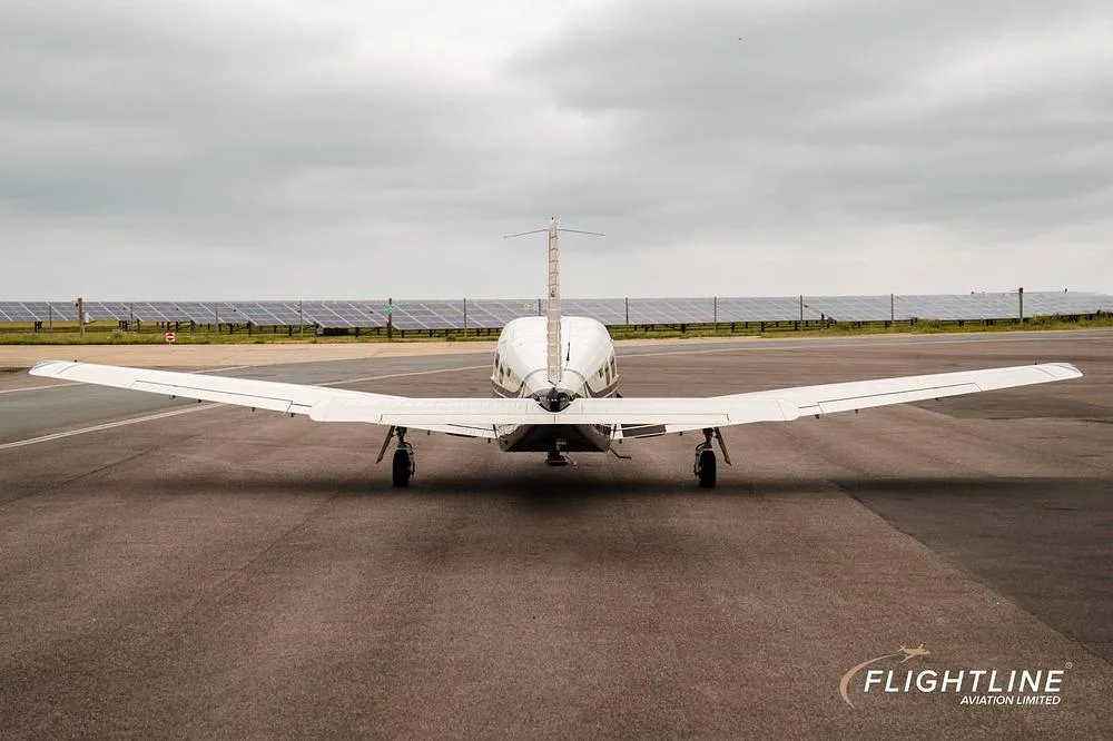 A rear view of a white and black Piper Saratoga II TC, registration N446SE, parked on an asphalt runway with solar panels in the background.