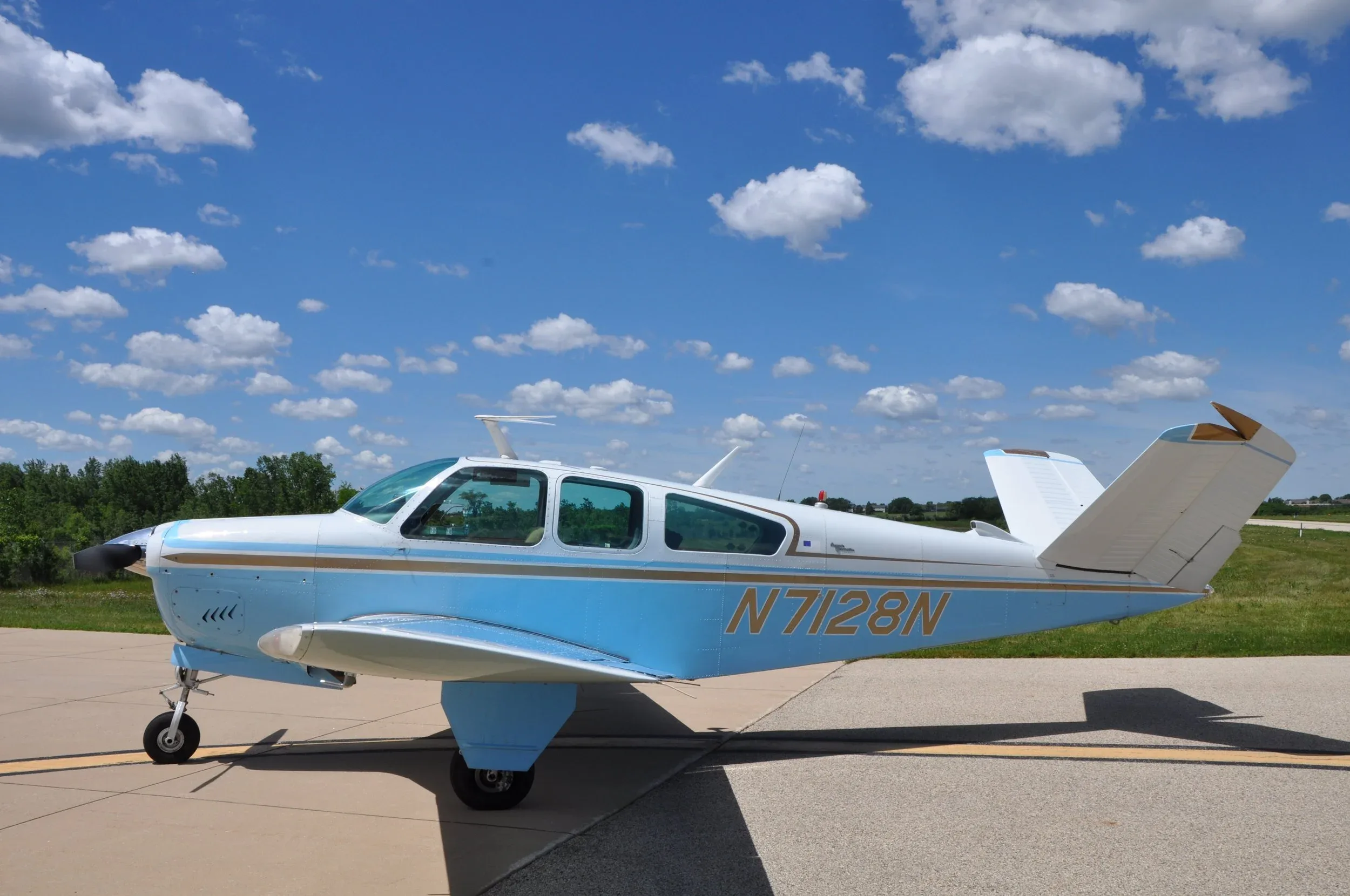 A side view of a light blue and white single-engine aircraft with a V-tail, showing the registration N7128N, parked on an asphalt runway.