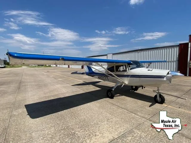 A white and blue Cessna 172G aircraft is parked on an airport apron under a blue sky.