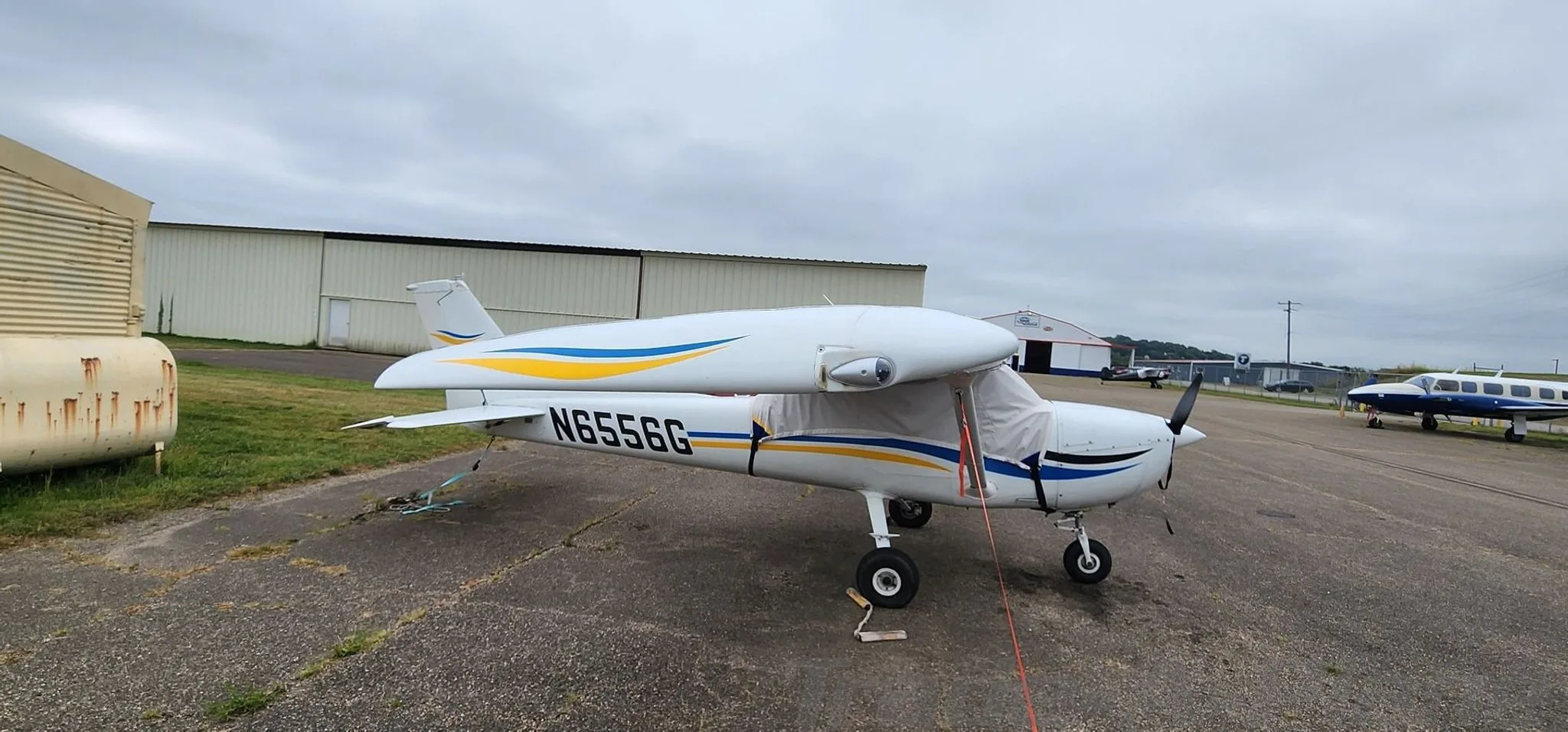A white Cessna 150 with blue and yellow stripes, covered with wing and cockpit covers, tied down on an airport tarmac.