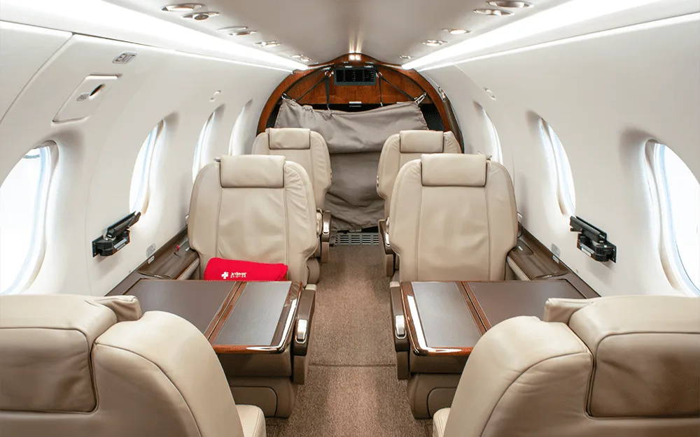 Interior view of a private aircraft cabin with beige leather seats and fold-out tables, looking towards the rear.