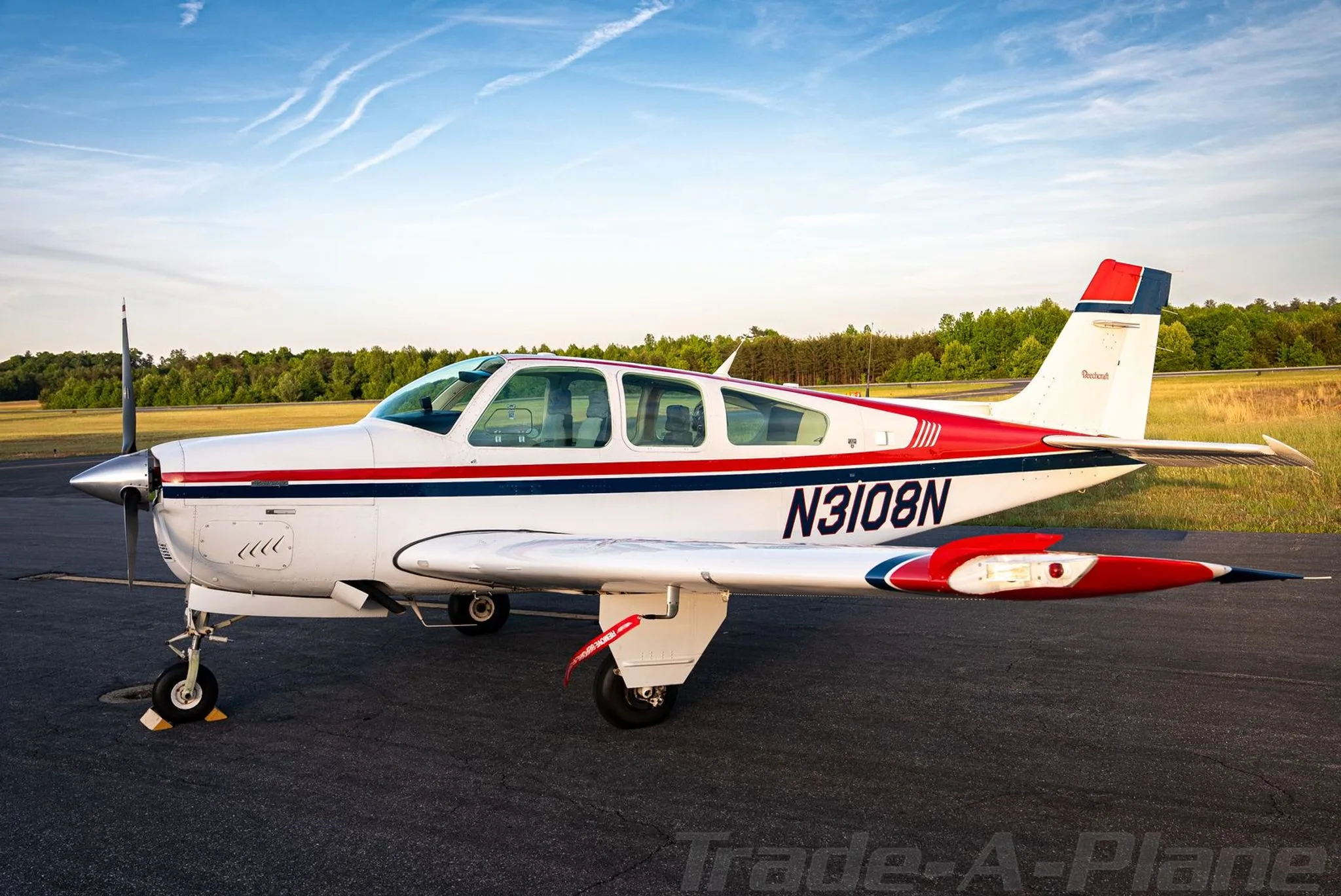 A white, red, and blue Beechcraft Bonanza N3108N parked on an asphalt ramp at sunset.