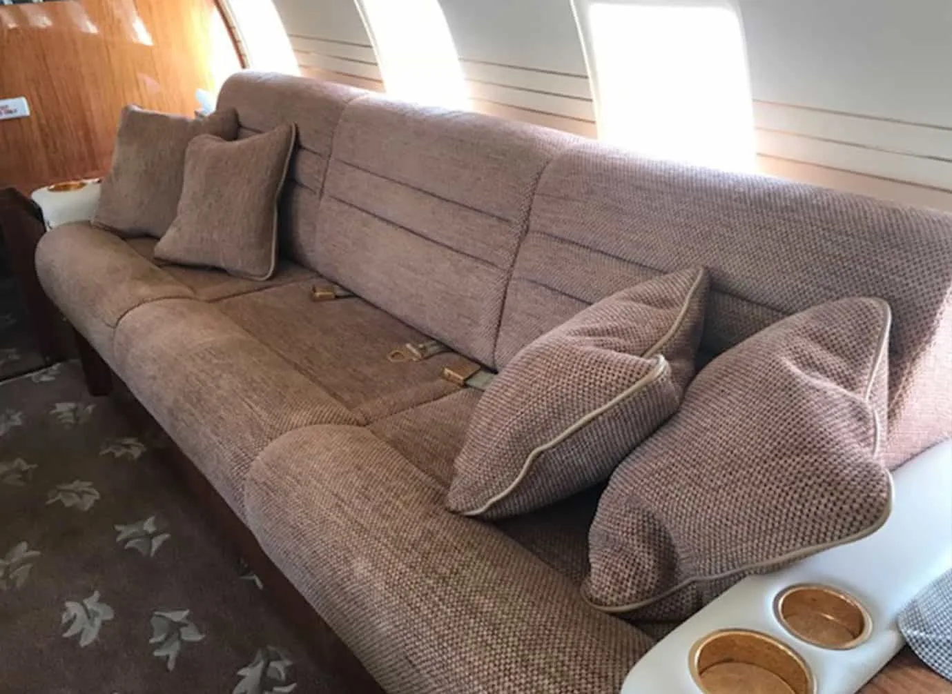 A brown fabric sofa with pillows and cup holders is seen inside an aircraft cabin.