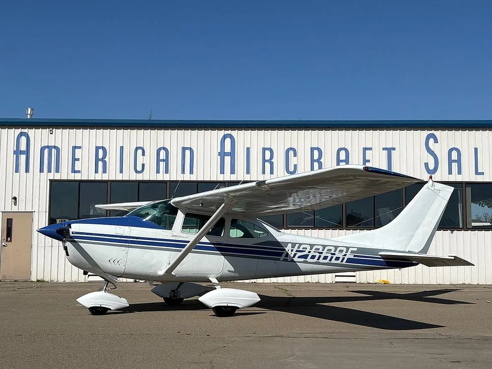 A white and blue Cessna 182 aircraft (N2668F) parked in front of an "American Aircraft Sales" hangar.