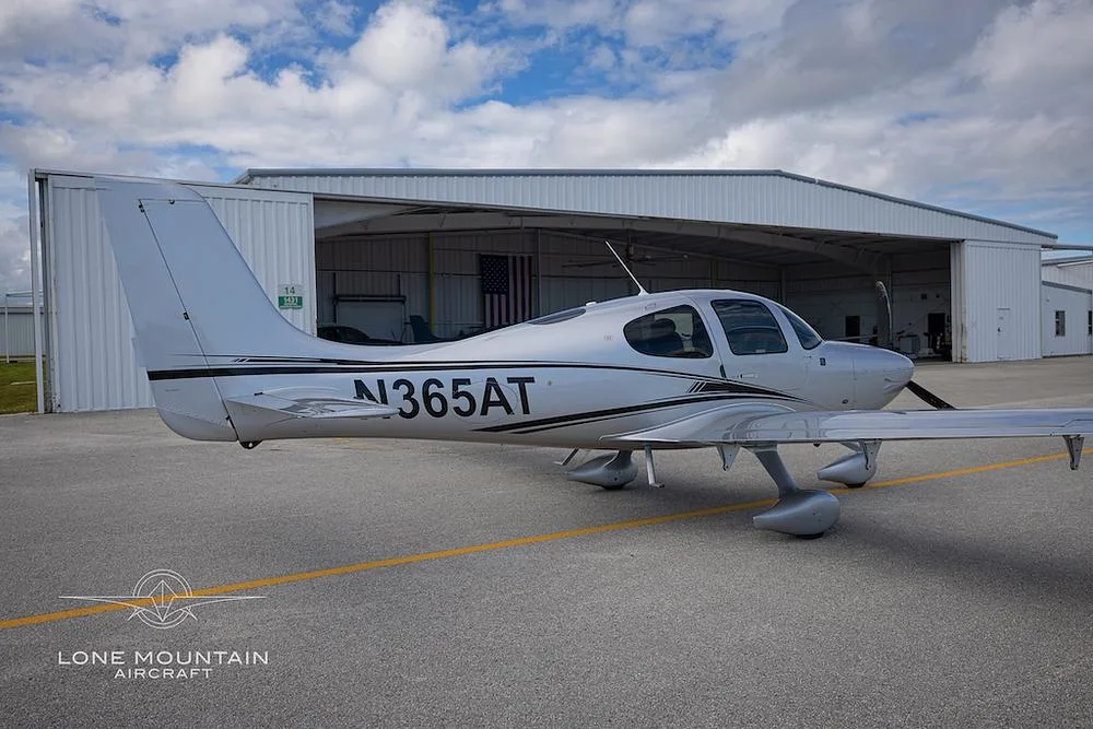 A silver Cirrus SR22 aircraft with black and white stripes, parked on the tarmac in front of an open hangar.