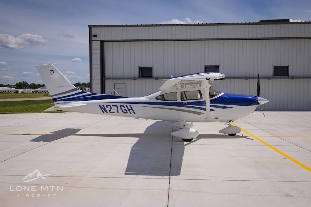 A side view of a white and blue Cessna 182 Skylane (N27GH) parked on an airport tarmac.