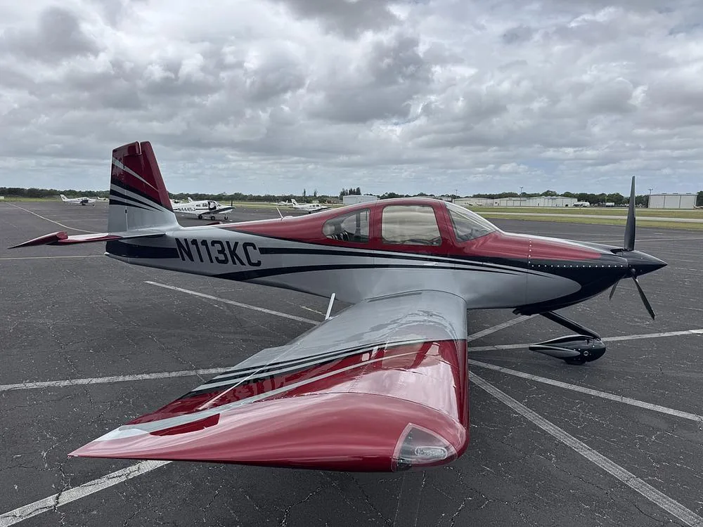 A red, silver, and black single-engine aircraft (N113KC) parked on an airport tarmac with other planes in the distance.