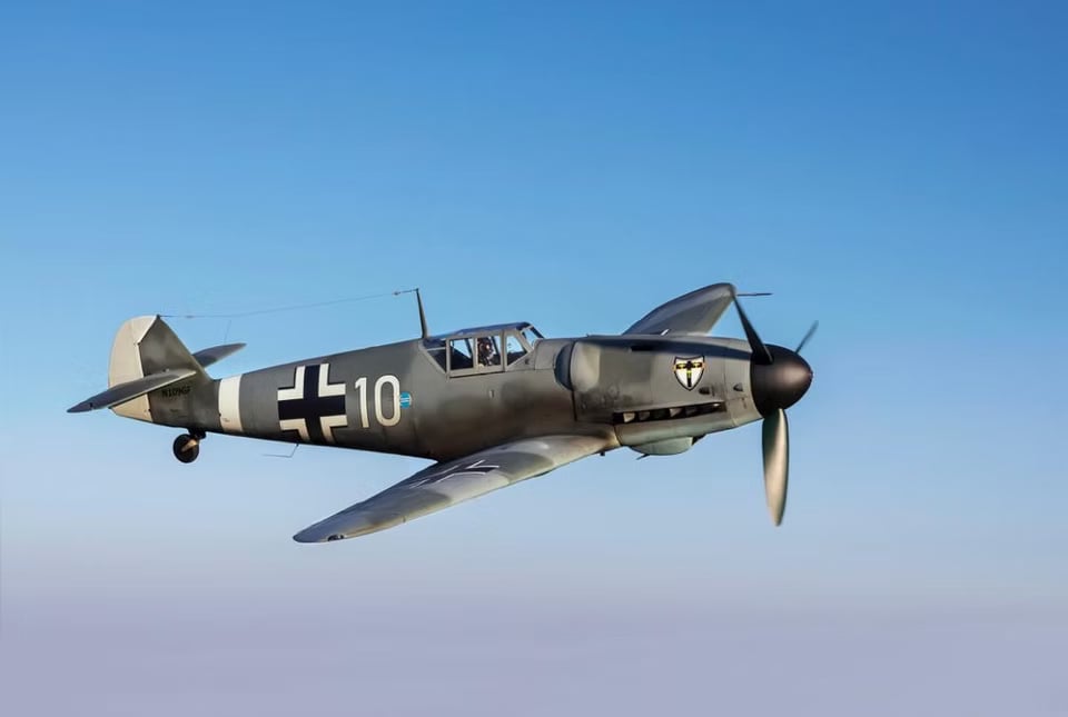 A gray Messerschmitt Bf 109 aircraft in flight over a forest.