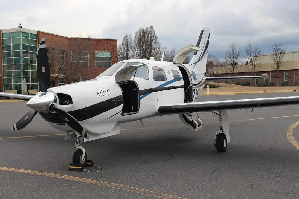A white Piper M350 aircraft with black and blue stripes, registration N377ST, parked on an asphalt tarmac with its main and cargo doors open.