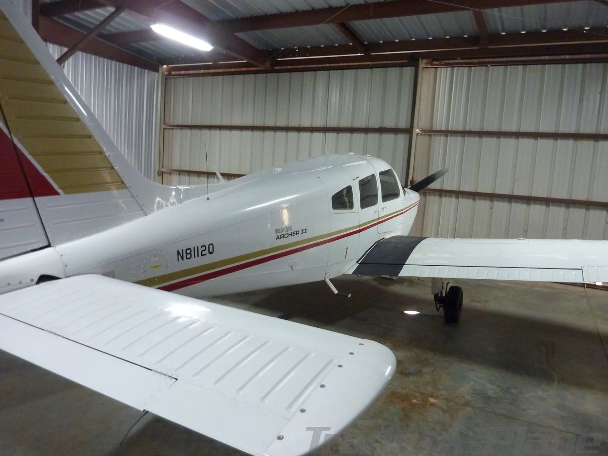 A top-down side view of a white Piper Archer II aircraft with red and gold stripes, registration N8112Q, in a hangar.