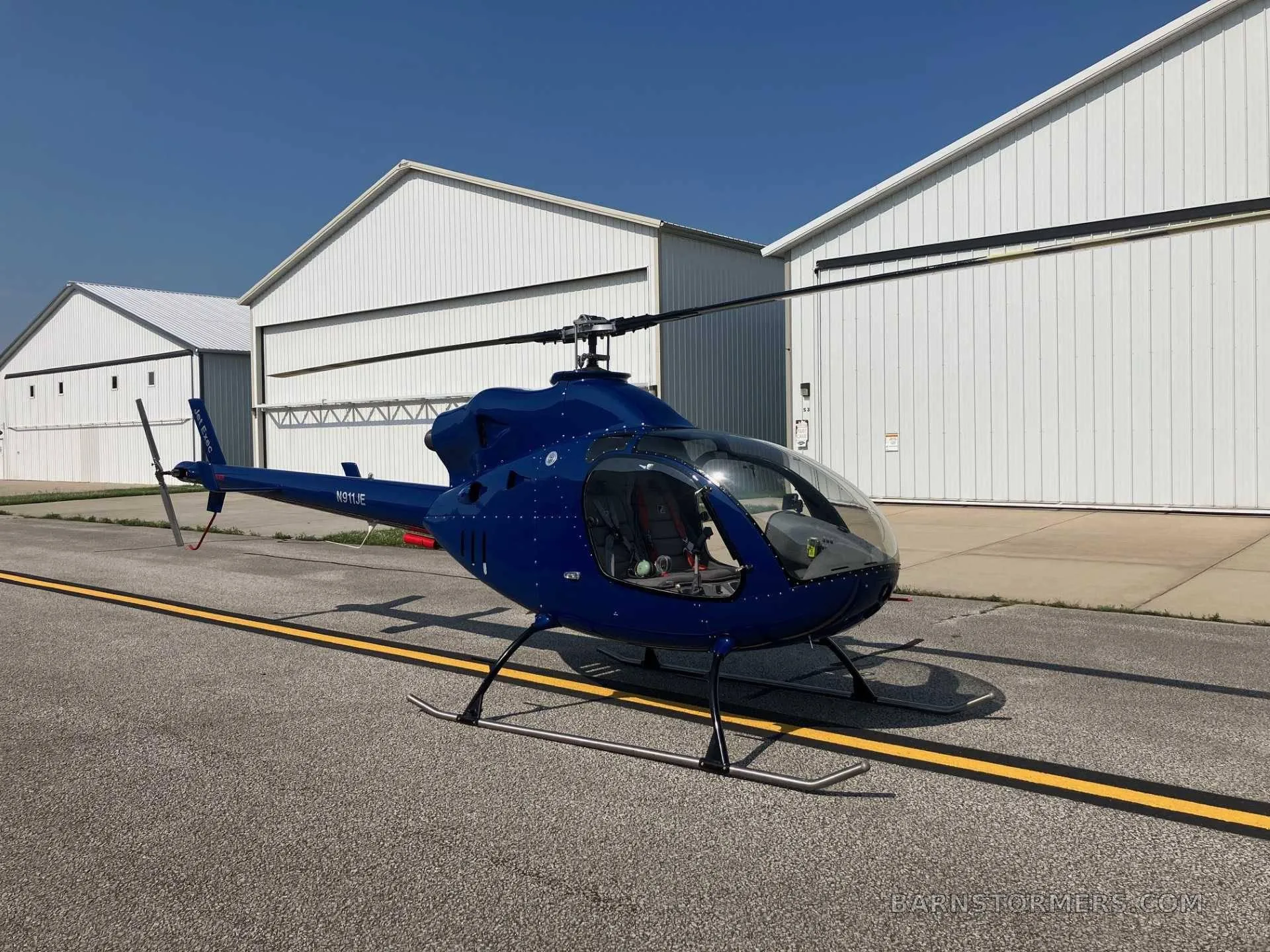 A blue helicopter (N911JE) with skids parked on an asphalt tarmac in front of white hangars under a clear sky.
