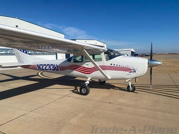 A white Cessna 172 with patriotic red and blue stripes is parked on an airport tarmac.