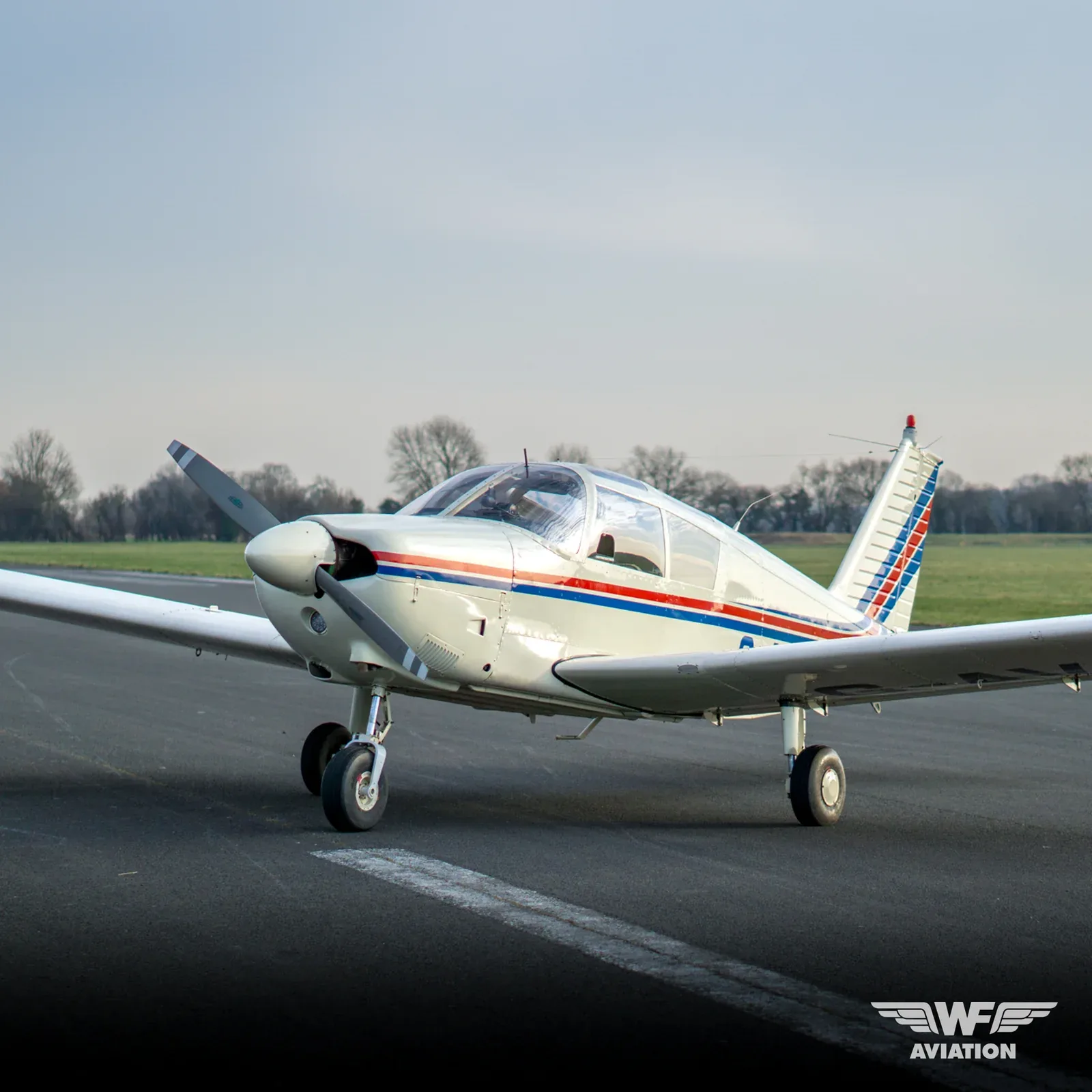 A white single-engine propeller aircraft with red and blue stripes on its fuselage and tail, parked on an airfield.