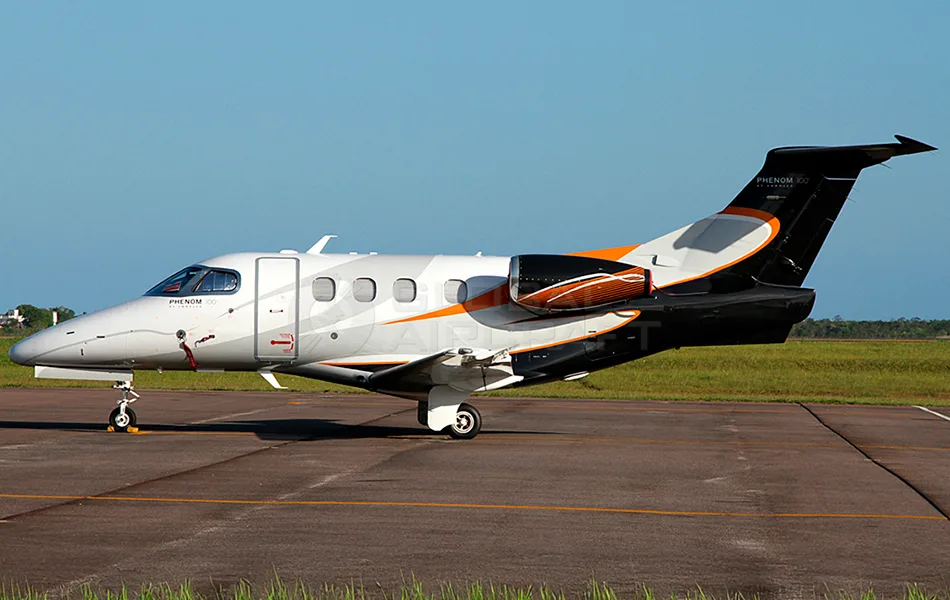 A white Embraer Phenom 100 business jet with black and orange livery parked on a tarmac under a clear sky.