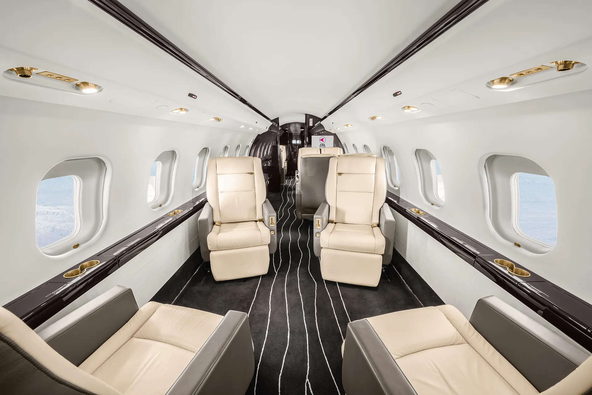 A wide-angle view of a private jet cabin interior with four cream leather seats arranged in two pairs facing each other, and a dark striped aisle.