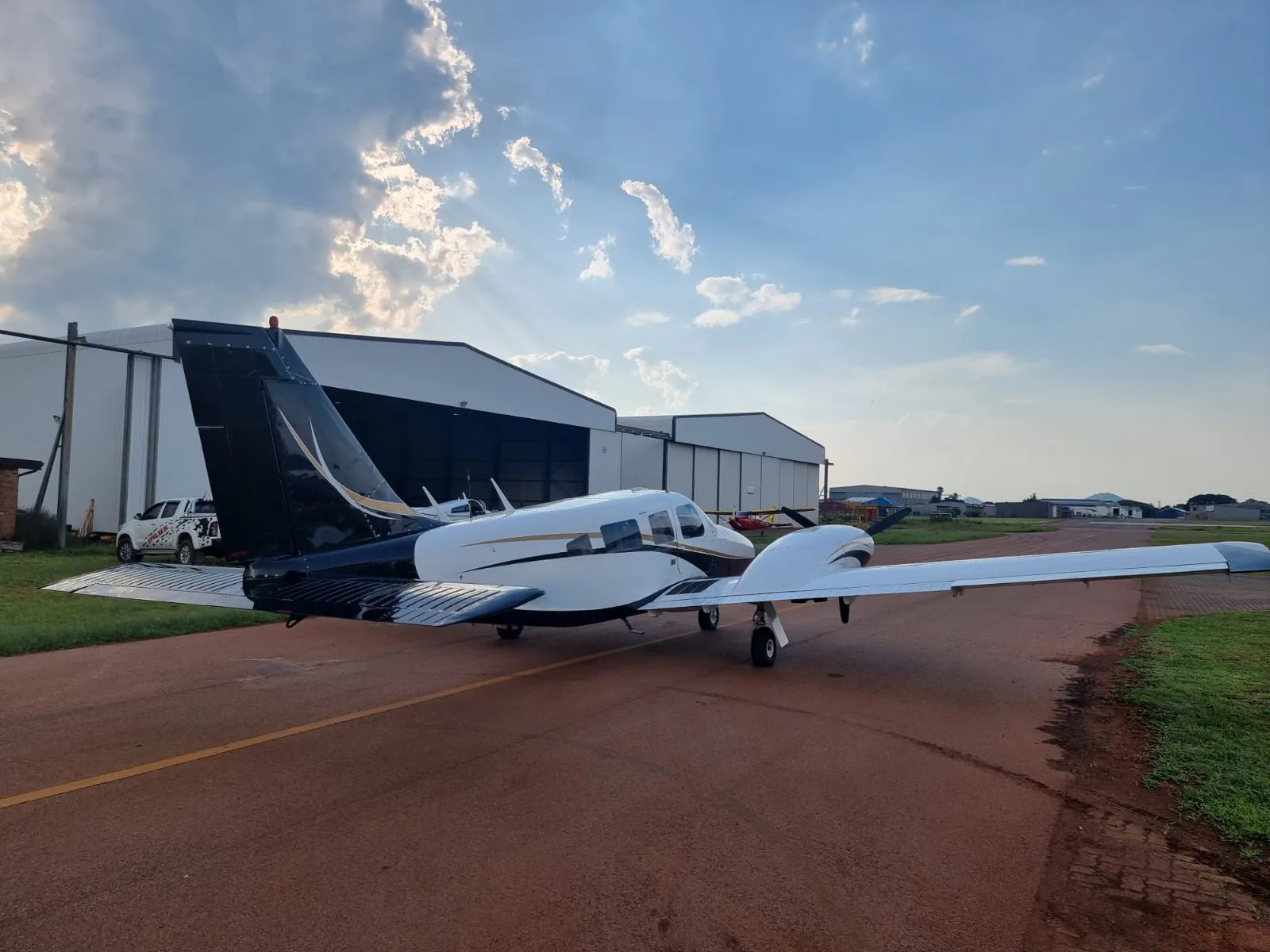 A white and black light twin-engine aircraft with gold accents is parked on a tarmac in front of hangars under a partly cloudy sky.