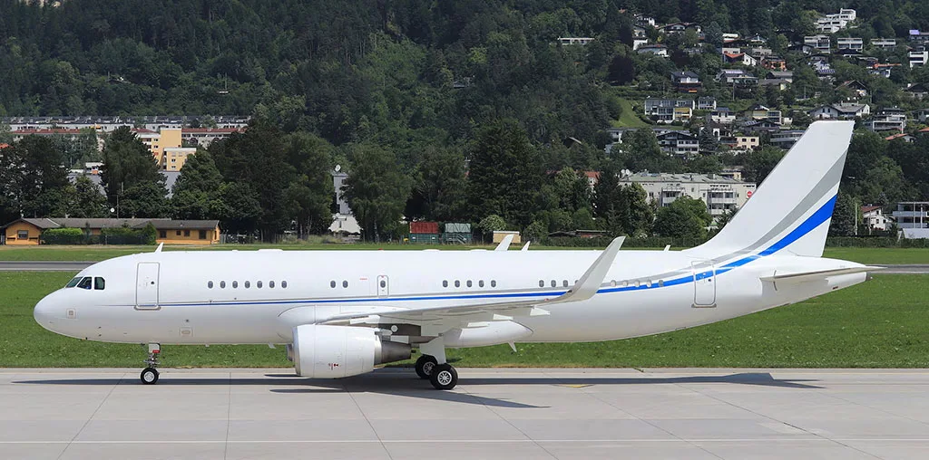 A white Airbus A320 family aircraft with blue and grey stripes on the tail and fuselage, parked on an tarmac with a green hillside and town in the background.