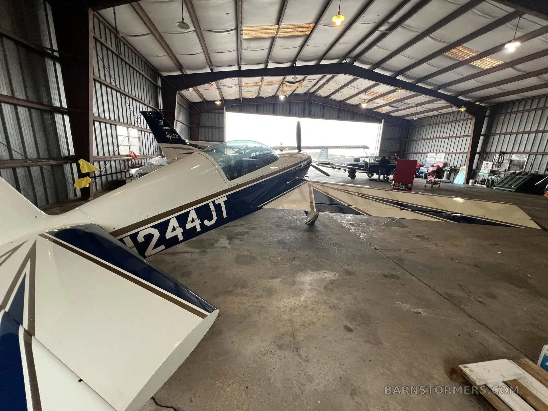A white and blue Extra 300L aircraft with tail number N244JT is parked inside a metal hangar, viewed from the rear-left.