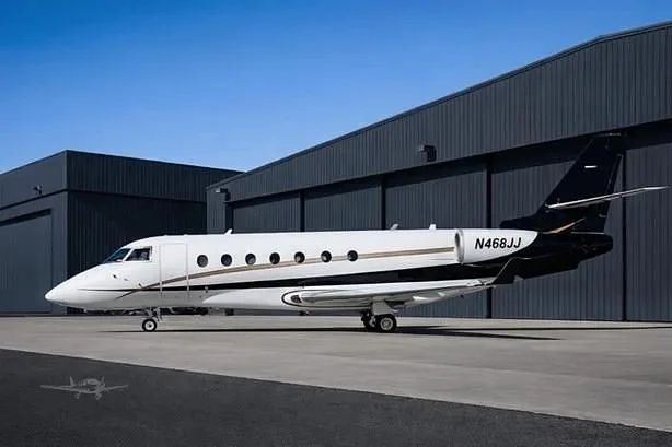 A white and black Gulfstream G200 private jet, registration N468JJ, parked on an tarmac in front of dark hangars.
