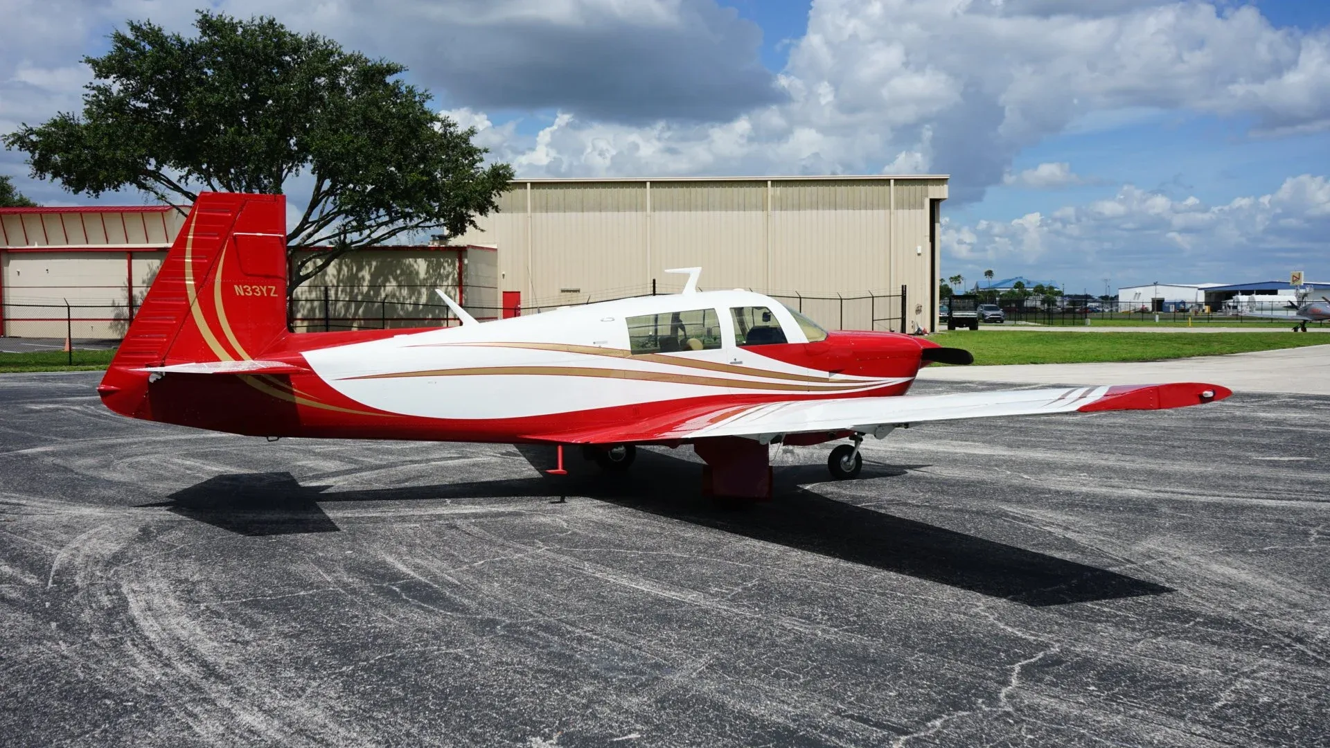 A red and white single-engine aircraft with gold stripes parked on an asphalt apron next to a hangar.