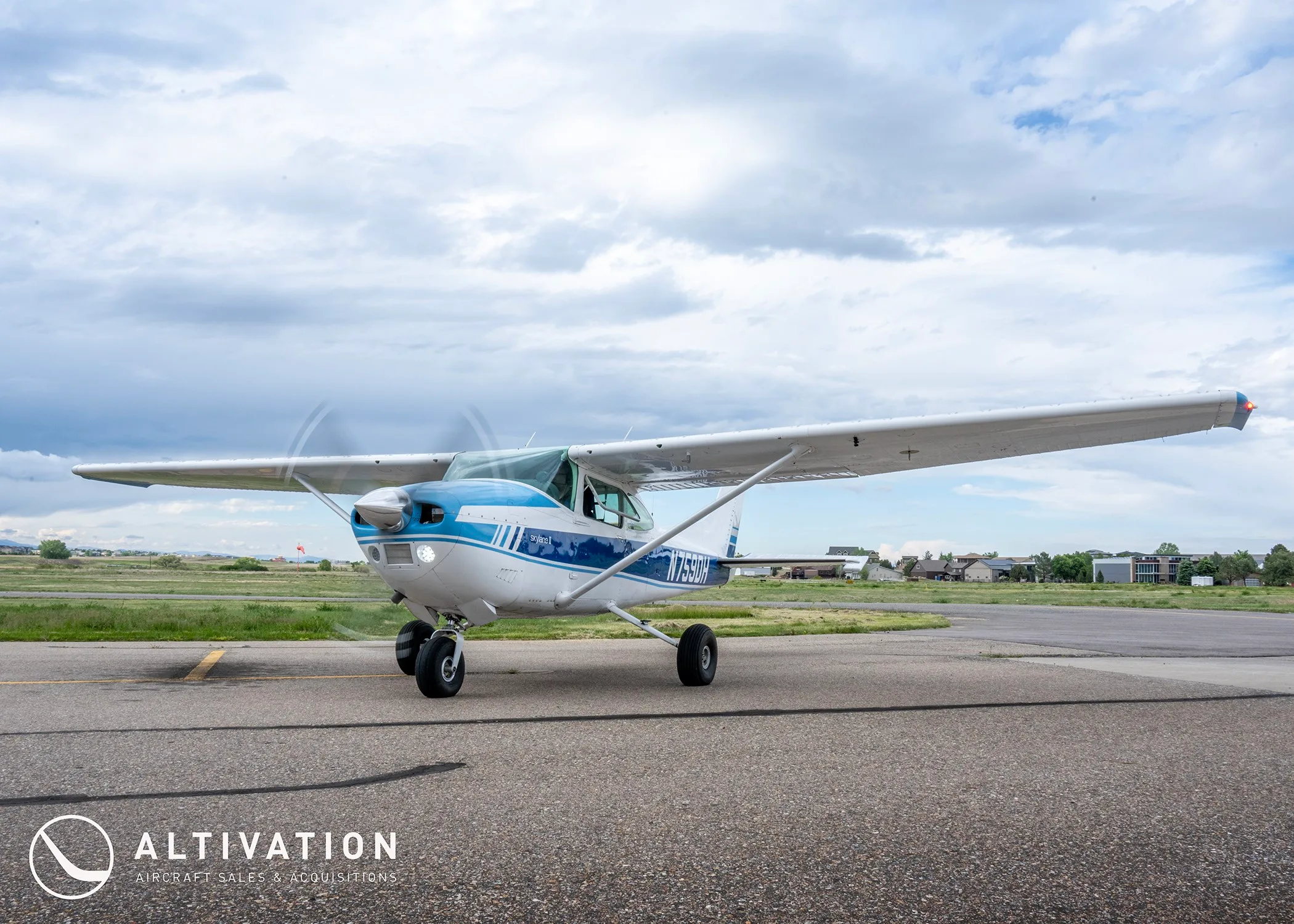 A white and blue Cessna 182 Skylane II (N759DH) with its propeller spinning, on an airfield under a cloudy sky.