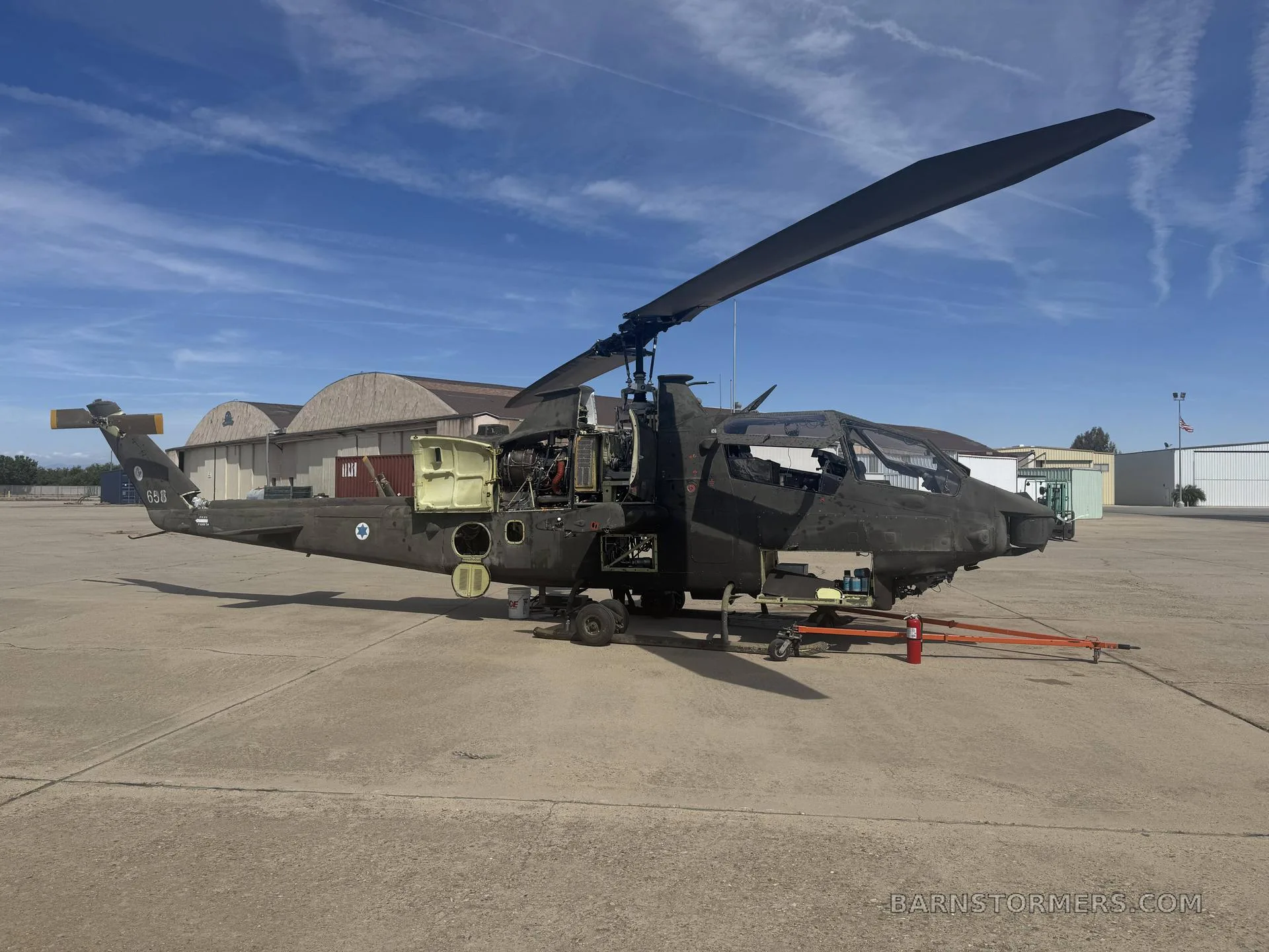 A partially disassembled dark green military helicopter, likely an Apache with Israeli markings, sits on a concrete airfield under a blue sky.