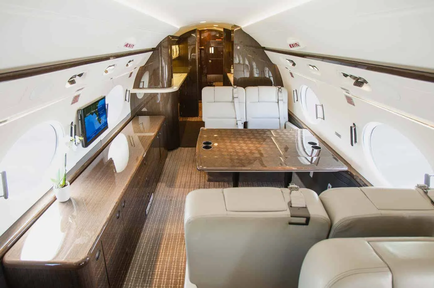 A luxurious aircraft cabin featuring a large dining table, light-colored seats, and dark wood cabinetry.