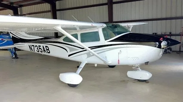 A white Cessna aircraft with black and grey stripes, registration N735AB, parked in a hangar.