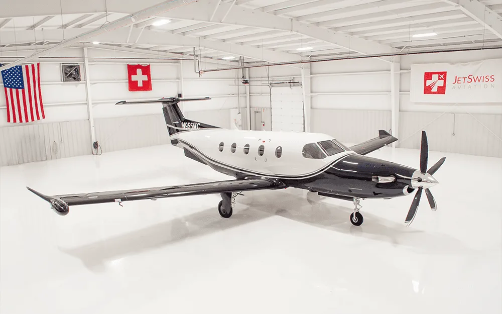 A black and white Pilatus PC-12 turboprop aircraft parked in a brightly lit hangar with US and Swiss flags.