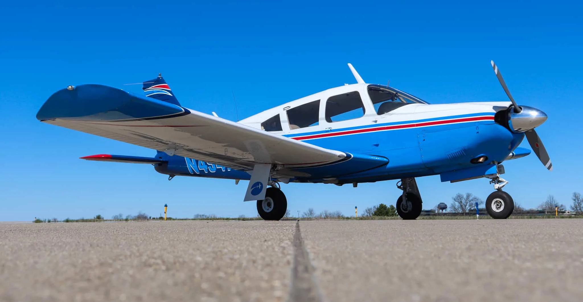 A low-angle side view of a blue and white Piper Cherokee aircraft with red stripes parked on an airfield.
