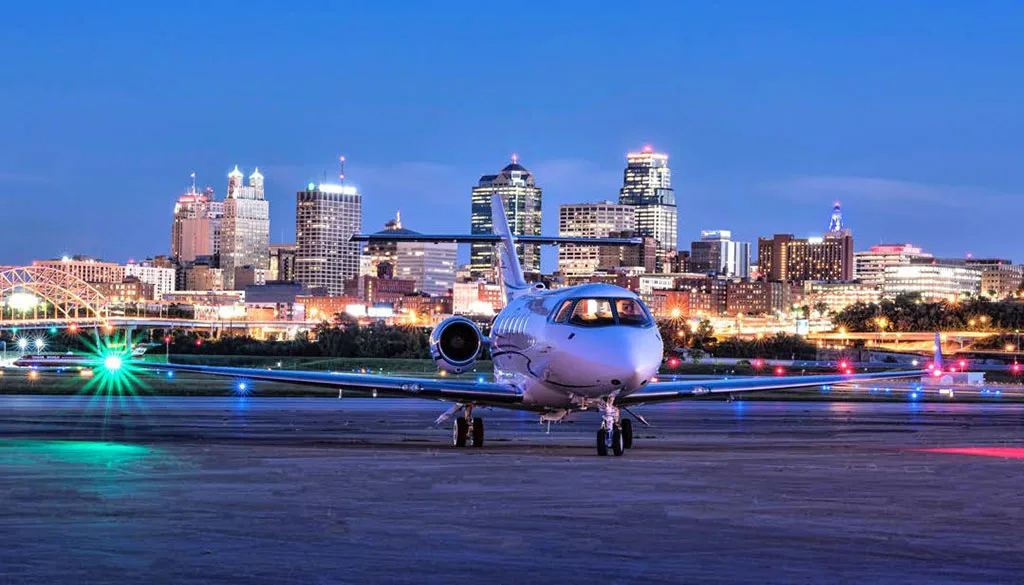 A white Hawker 900XP business jet is parked head-on on a tarmac at dusk, with the illuminated Kansas City skyline in the background.