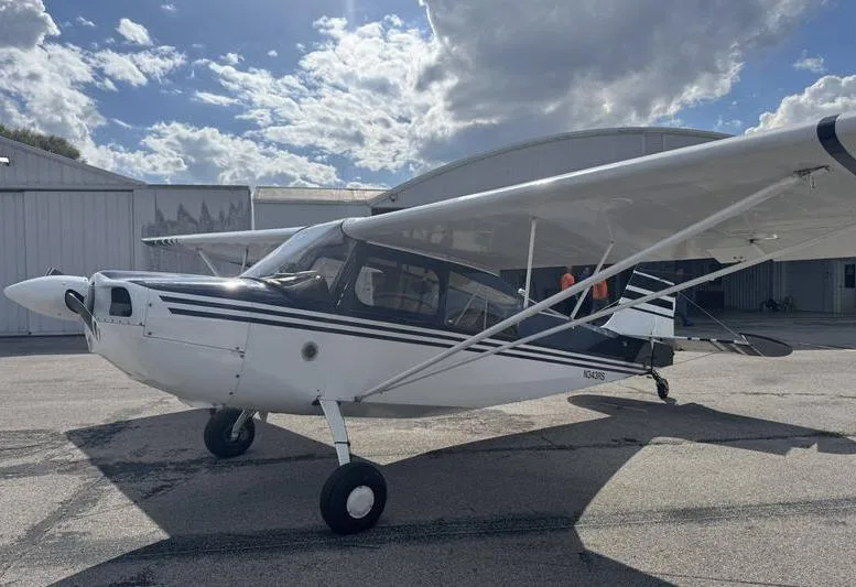 A white and dark blue high-wing aircraft, registration N343RS, parked on a tarmac with a hangar in the background under a partly cloudy sky.