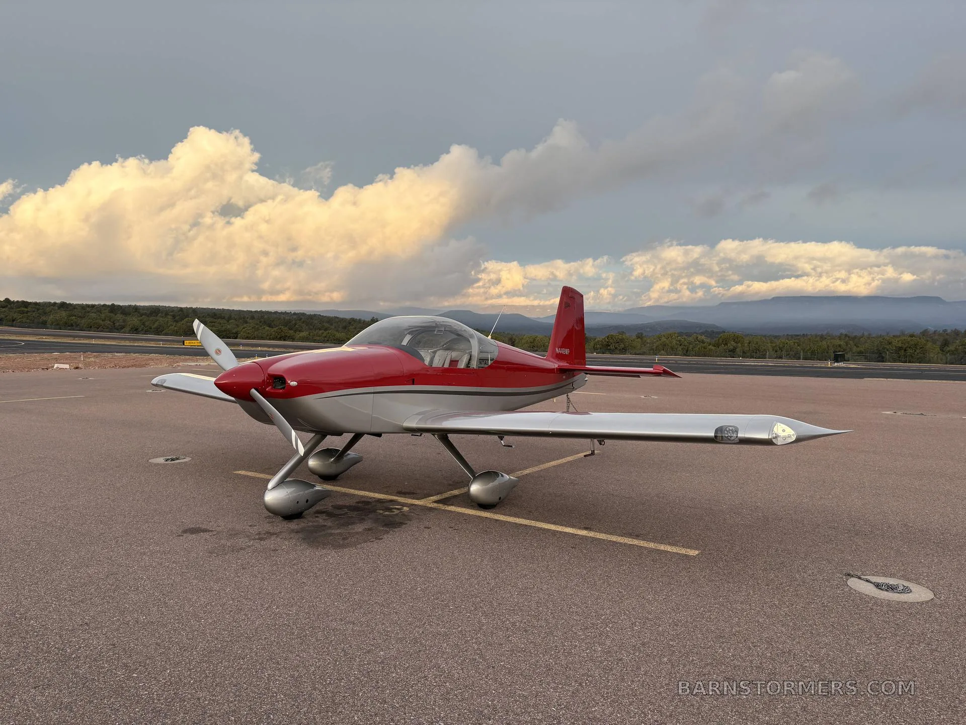 A red and silver single-engine aircraft, N448WP, parked on an airfield with mountains in the background under a dramatic cloudy sky.