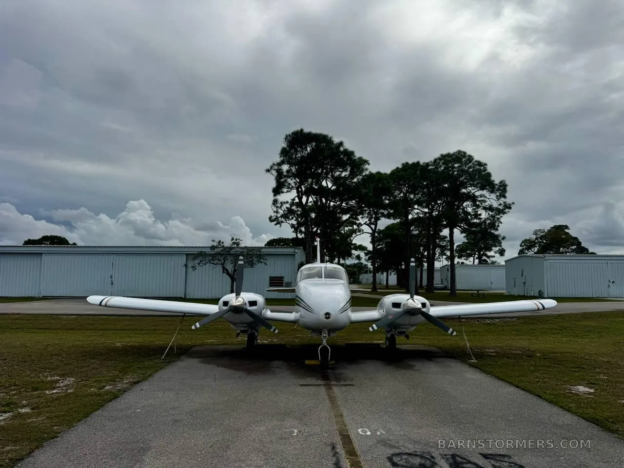 A head-on view of a white twin-engine aircraft with blue and gold stripes parked on an asphalt apron, under a cloudy sky.