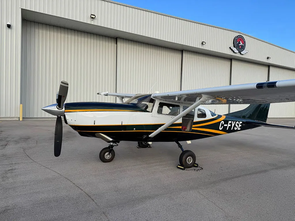 A dark green and white Cessna 182 with yellow stripes, registration C-FYSE, parked on an airfield in front of a hangar.