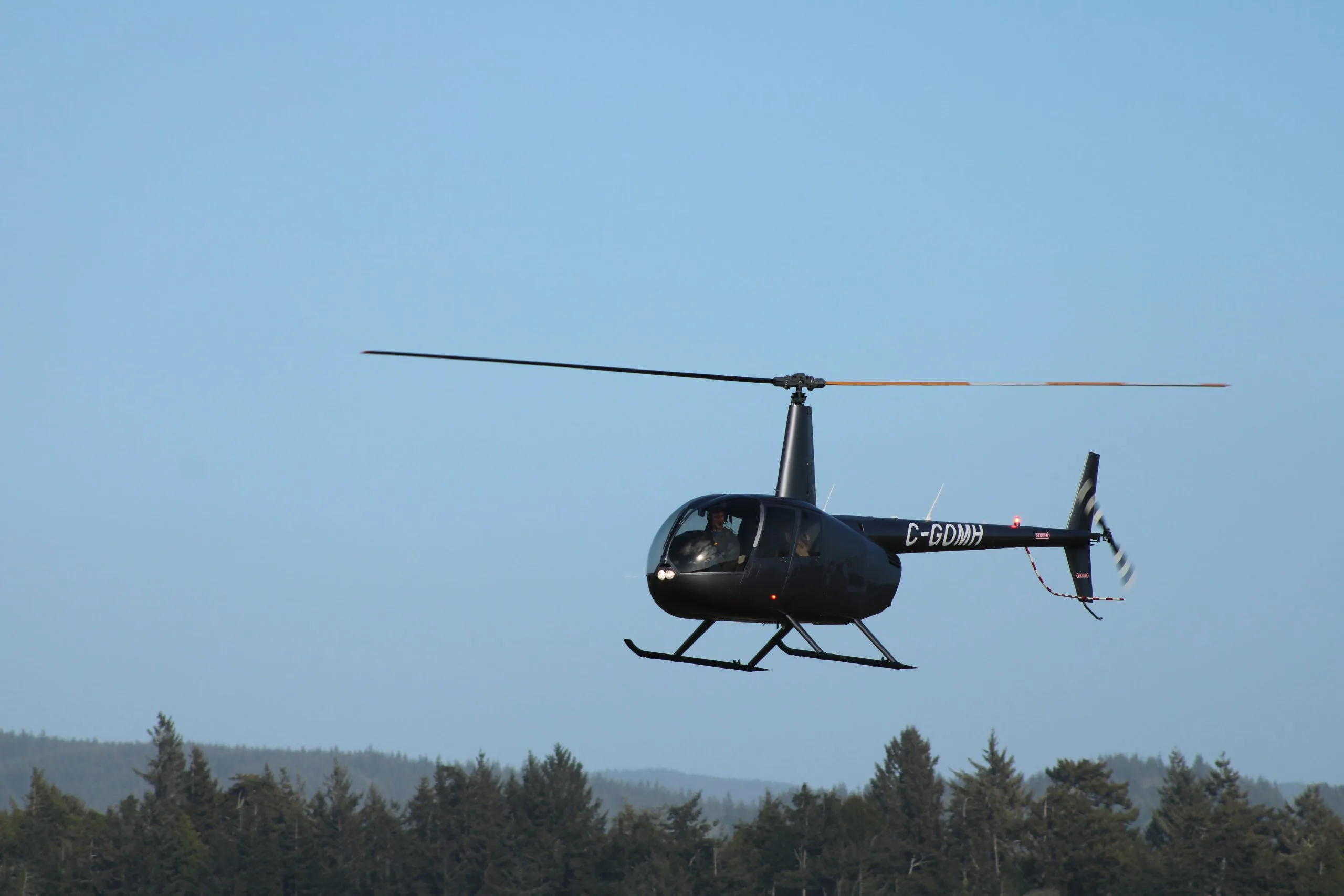 A dark grey Robinson R44 Raven II helicopter with registration C-GDMH flying low over a forest against a clear blue sky.