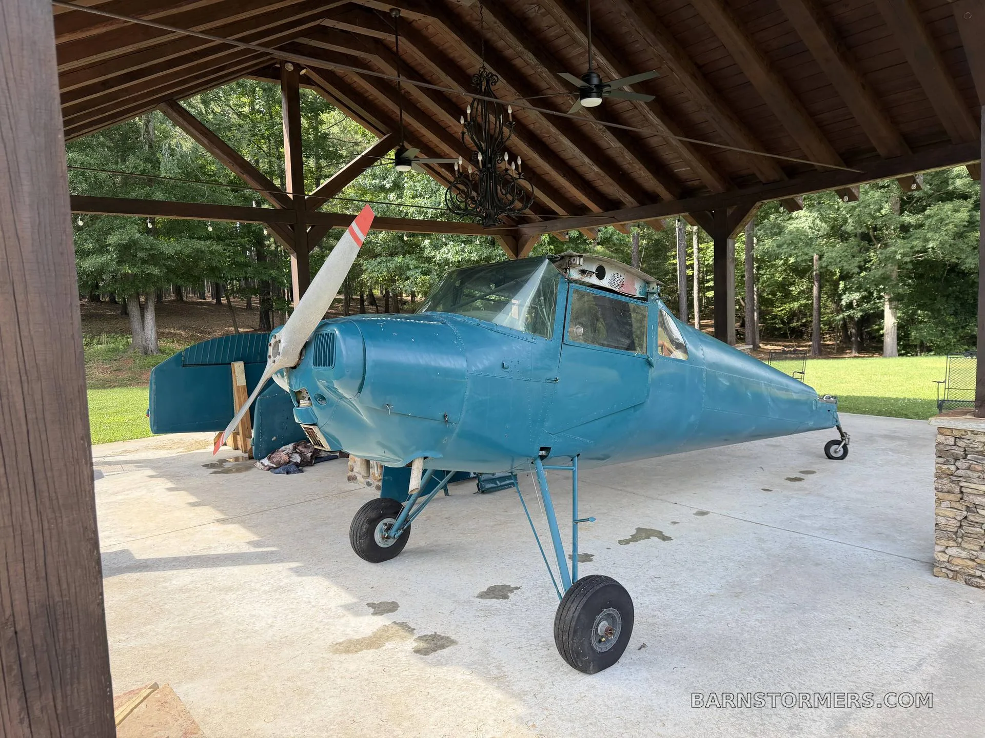 A teal aircraft fuselage with a propeller and landing gear, missing wings, parked under a wooden shelter.