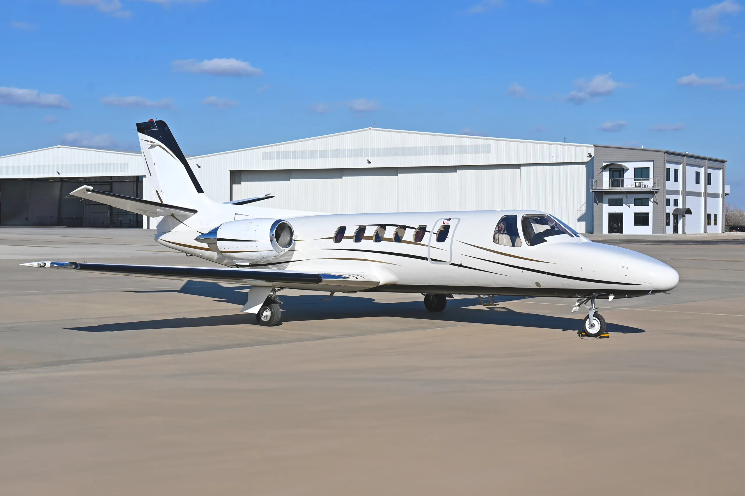 A white business jet with black and gold stripes parked on an airfield tarmac with hangars in the background.
