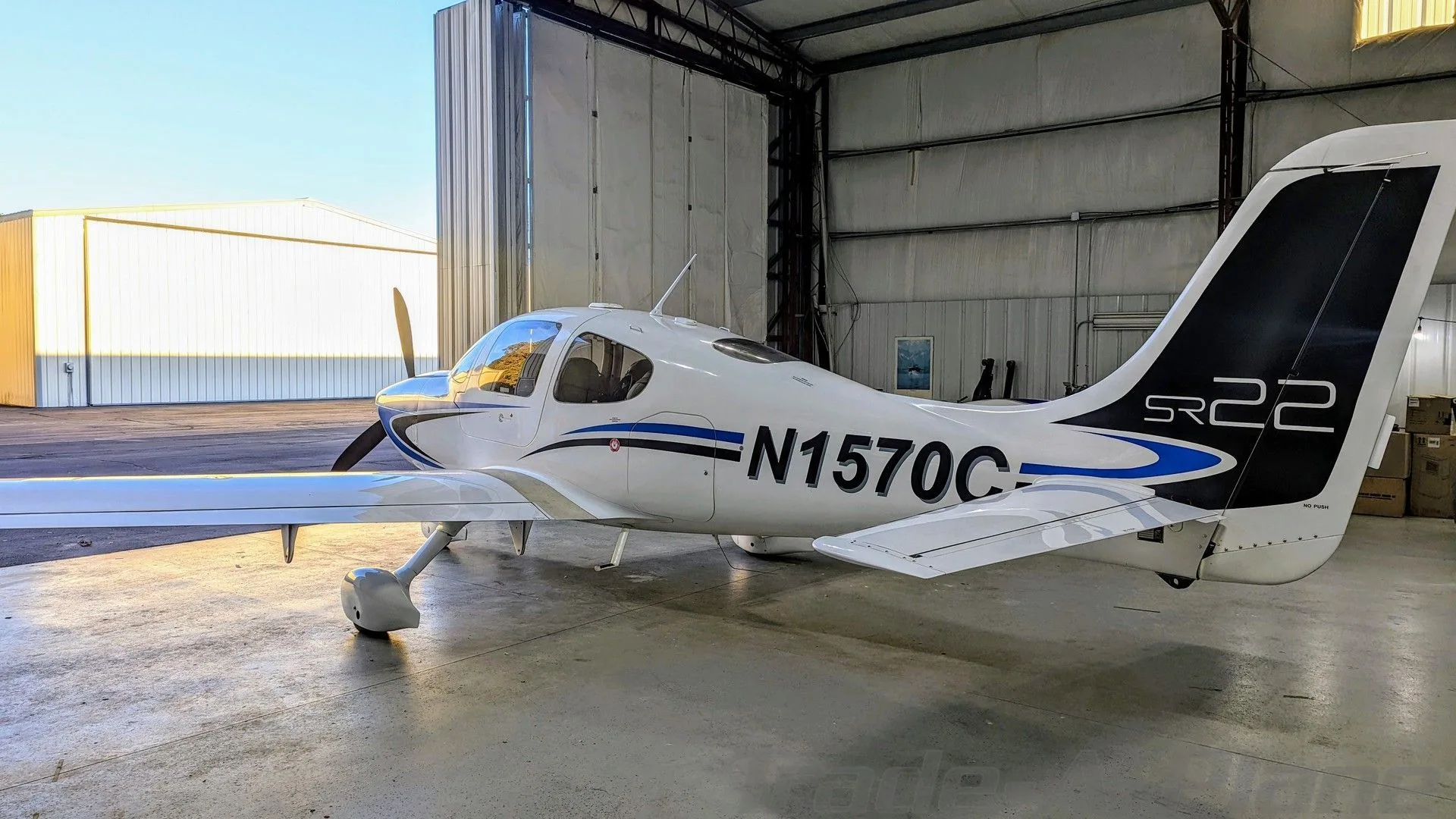 A white Cirrus SR22 aircraft with blue and black stripes, registration N1570C, parked inside a hangar, viewed from the rear.
