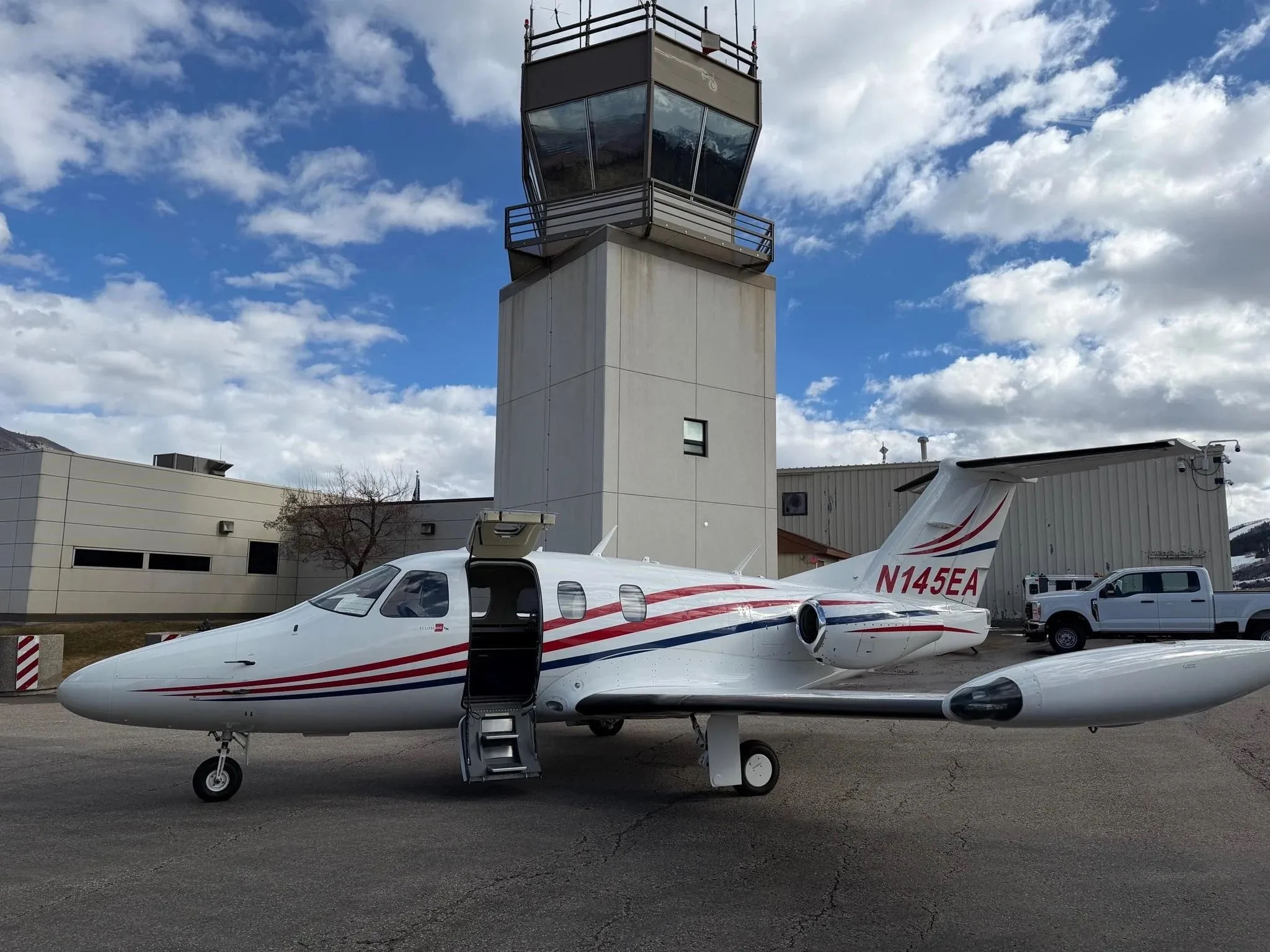 A white Eclipse jet with red and blue stripes, registration N145EA, parked on an airfield with an open door and a control tower in the background.