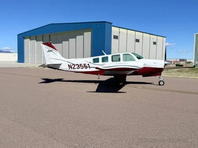 A white and red Beechcraft Bonanza aircraft is parked on an asphalt runway next to a blue and white hangar.