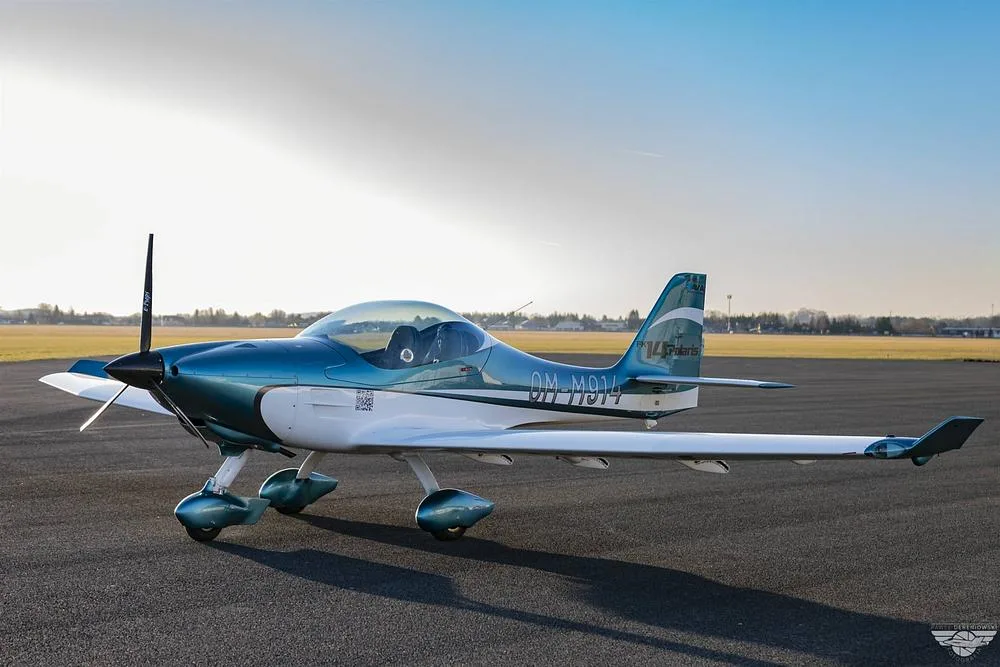A blue and white FK14 Polaris aircraft with registration OM-M914 parked on an asphalt runway at sunset.