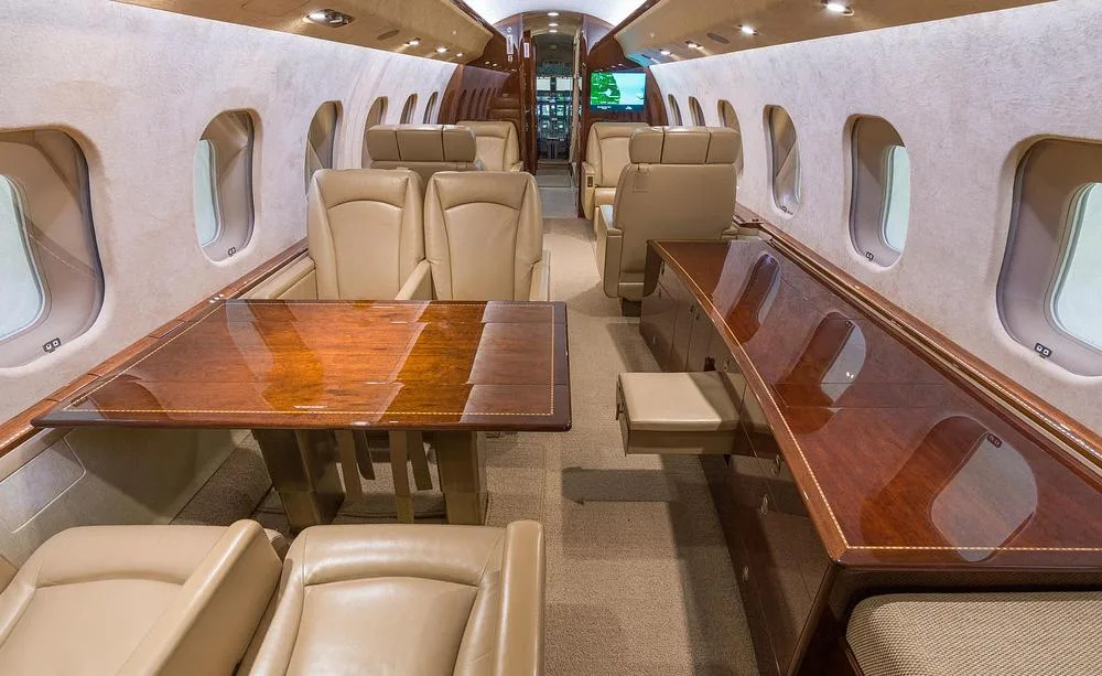 A view of the business jet's cabin interior, featuring a large wooden table and comfortable beige leather seats.