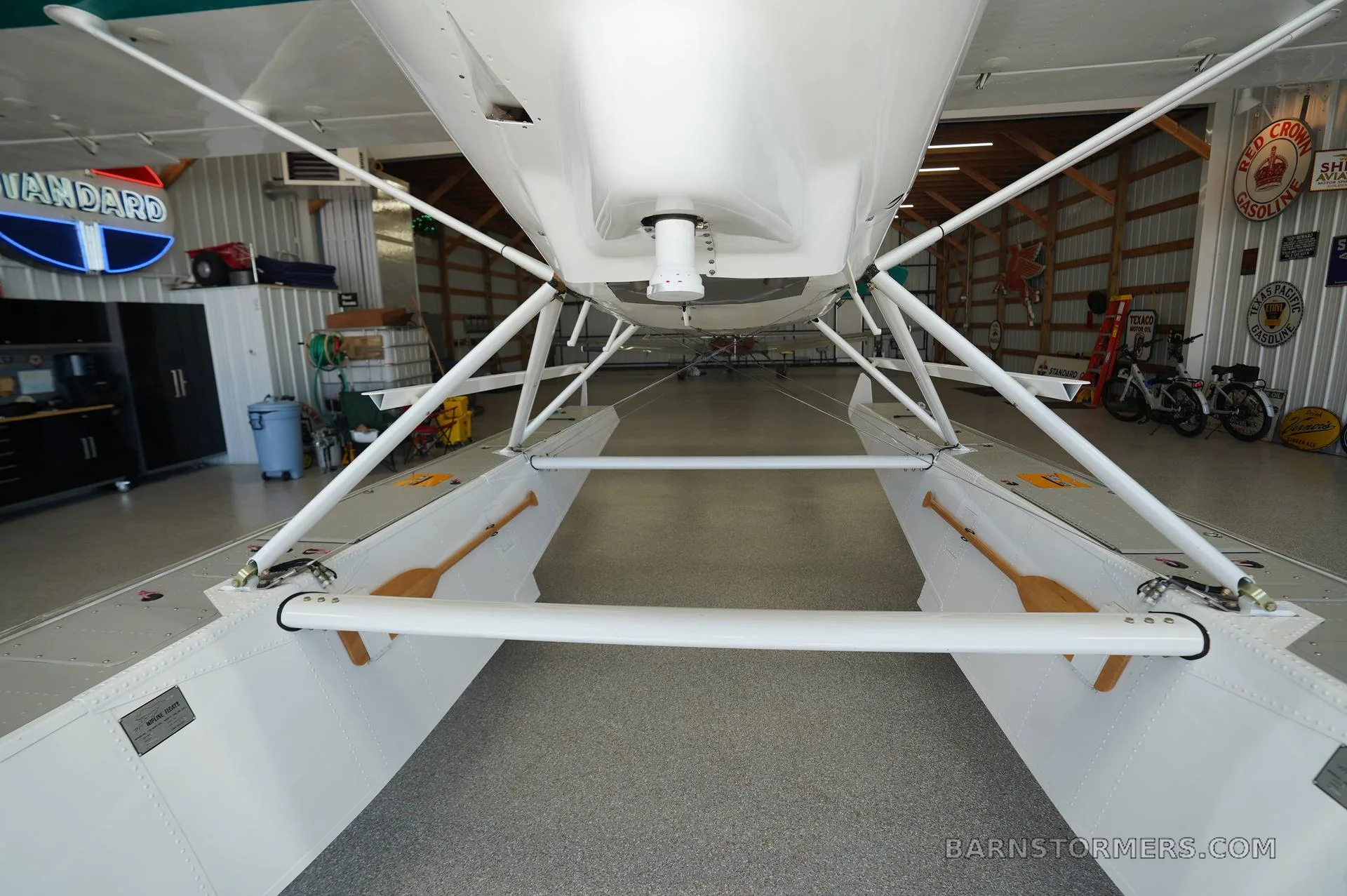 A close-up view of the white floats and underside of a floatplane, showing two wooden paddles stored on the floats, inside a hangar.