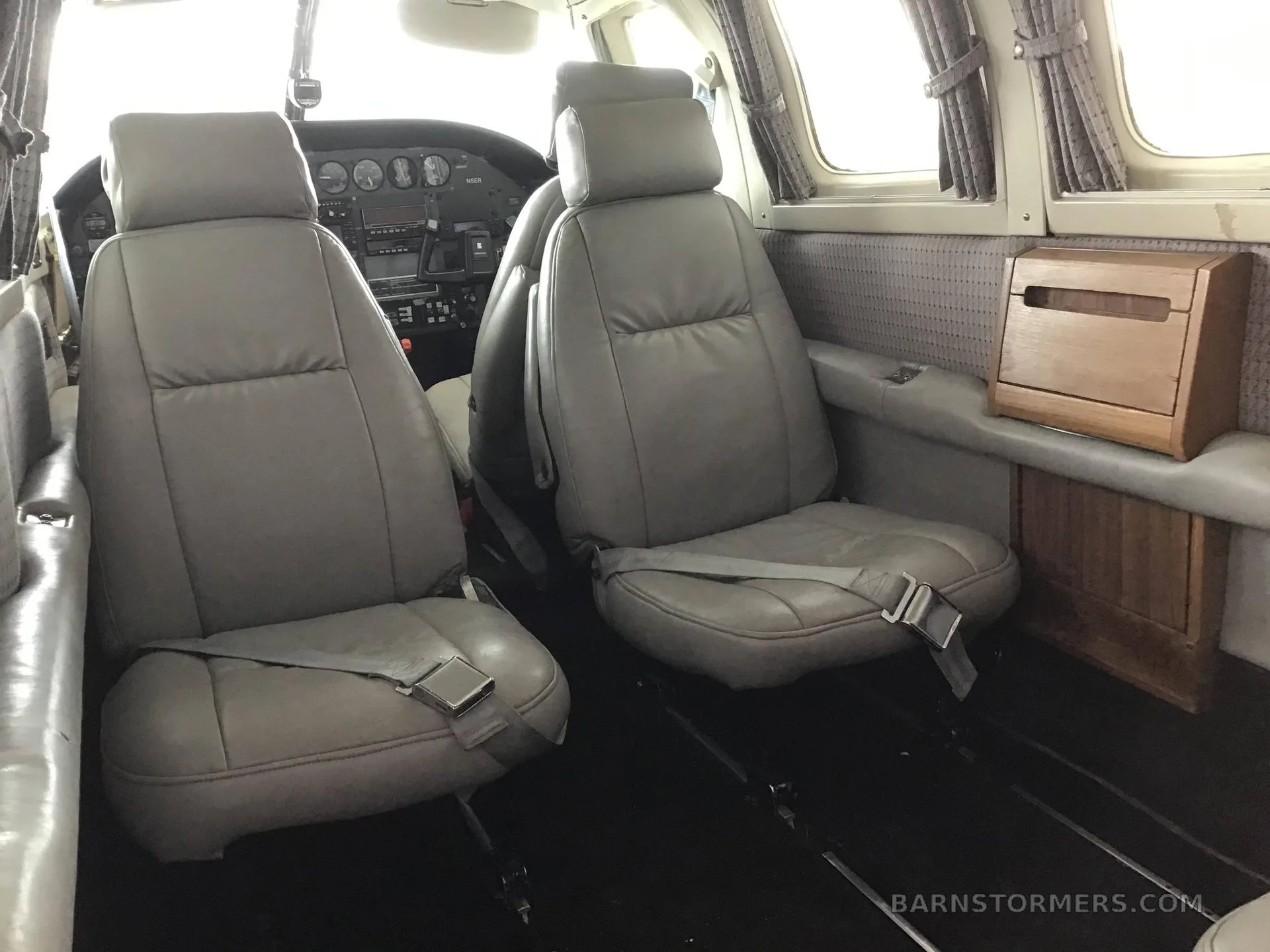The interior cabin of an aircraft, showing two grey leather passenger seats and a wooden tissue box holder.